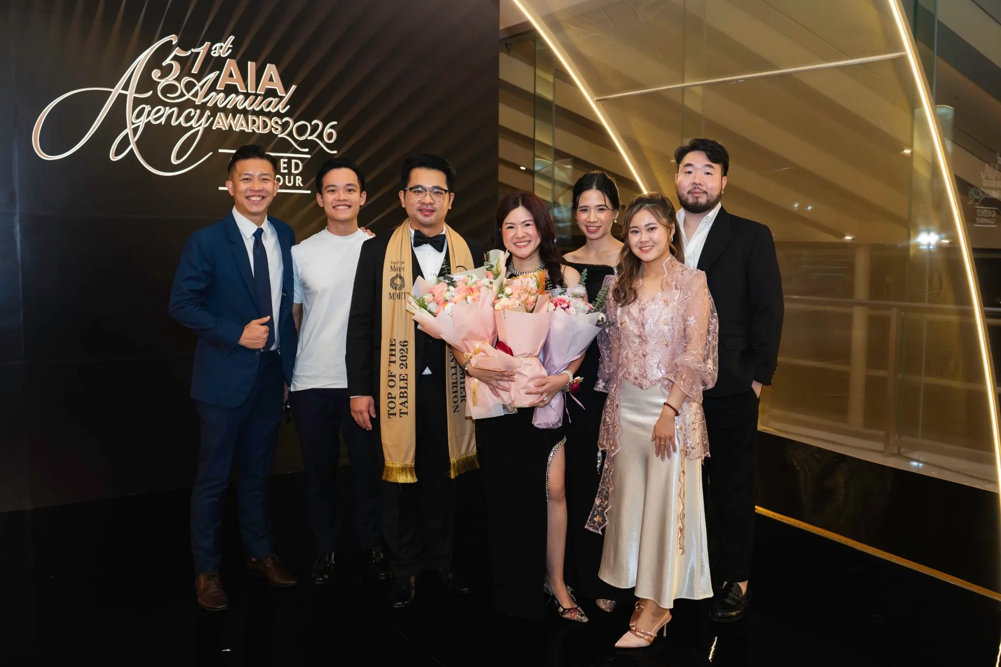 Shermene holding a bouquet, standing with six other people in front of the 51st AIA Annual Agency Awards 2026 sign