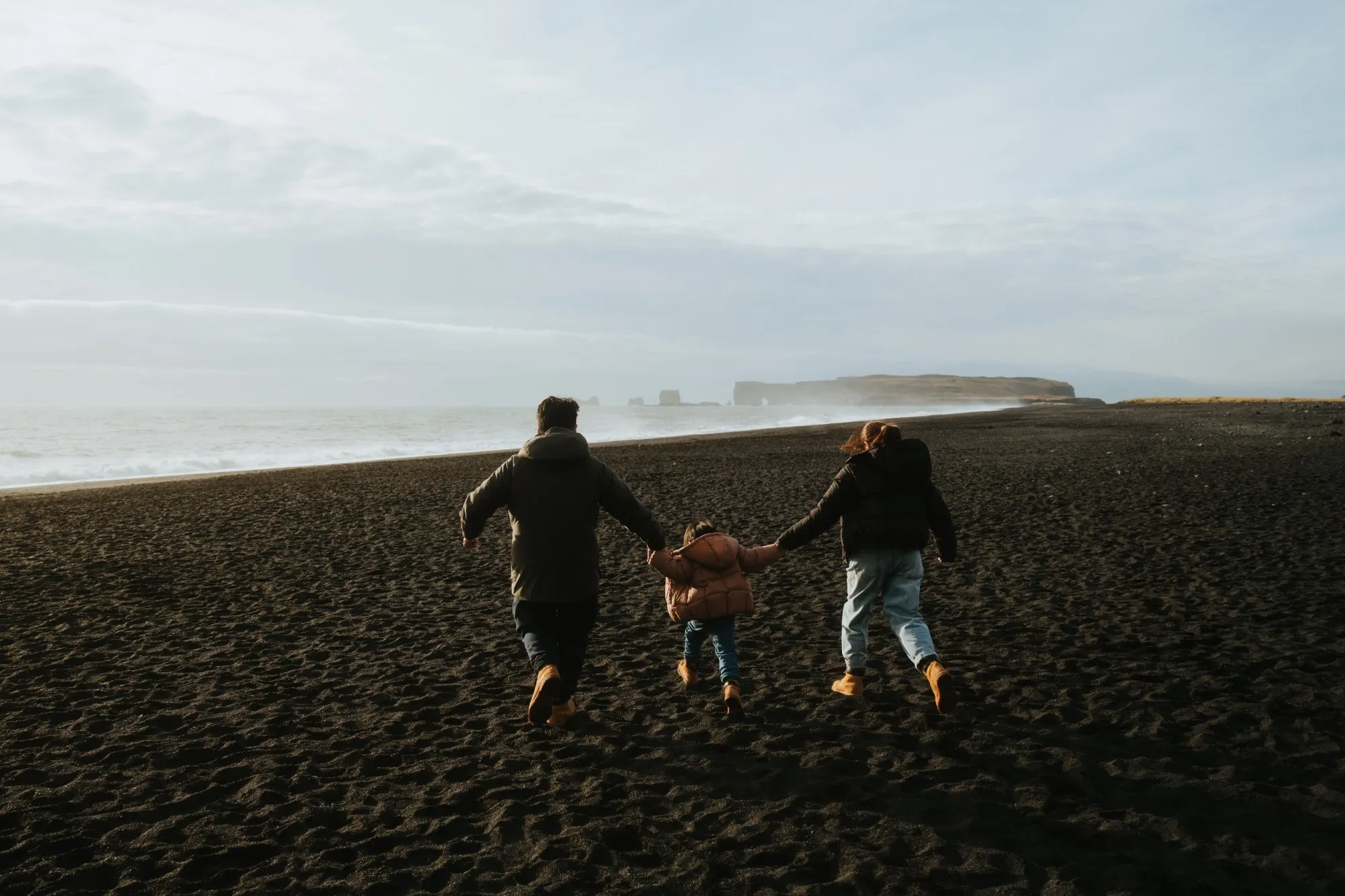 A man, a small child and a woman, seen from behind, running hand in hand across a wide black-sand beach toward the sea, with cliffs in the distance