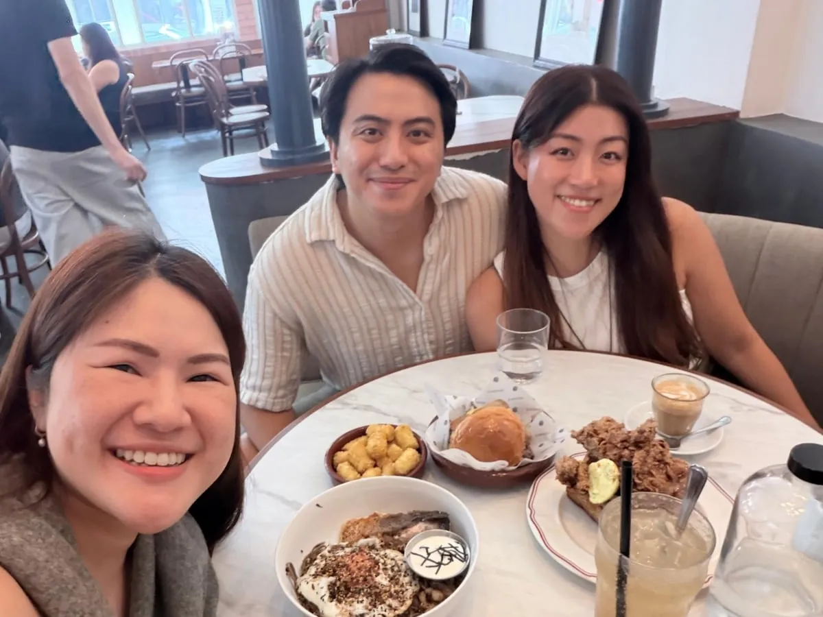 A selfie of three people smiling together at a cafe table with plates of food and drinks