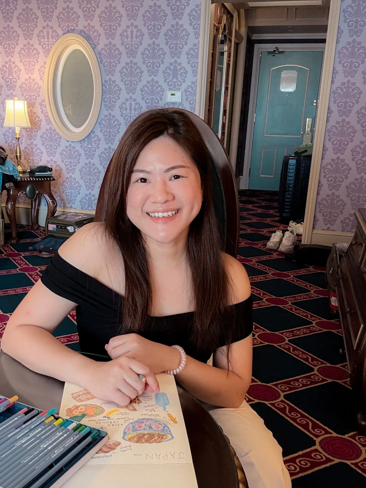 Shermene smiling at a table in a hotel room with a colored pencil in her hand, a Japan sketch of food on the page in front of her and a box of pencils beside it