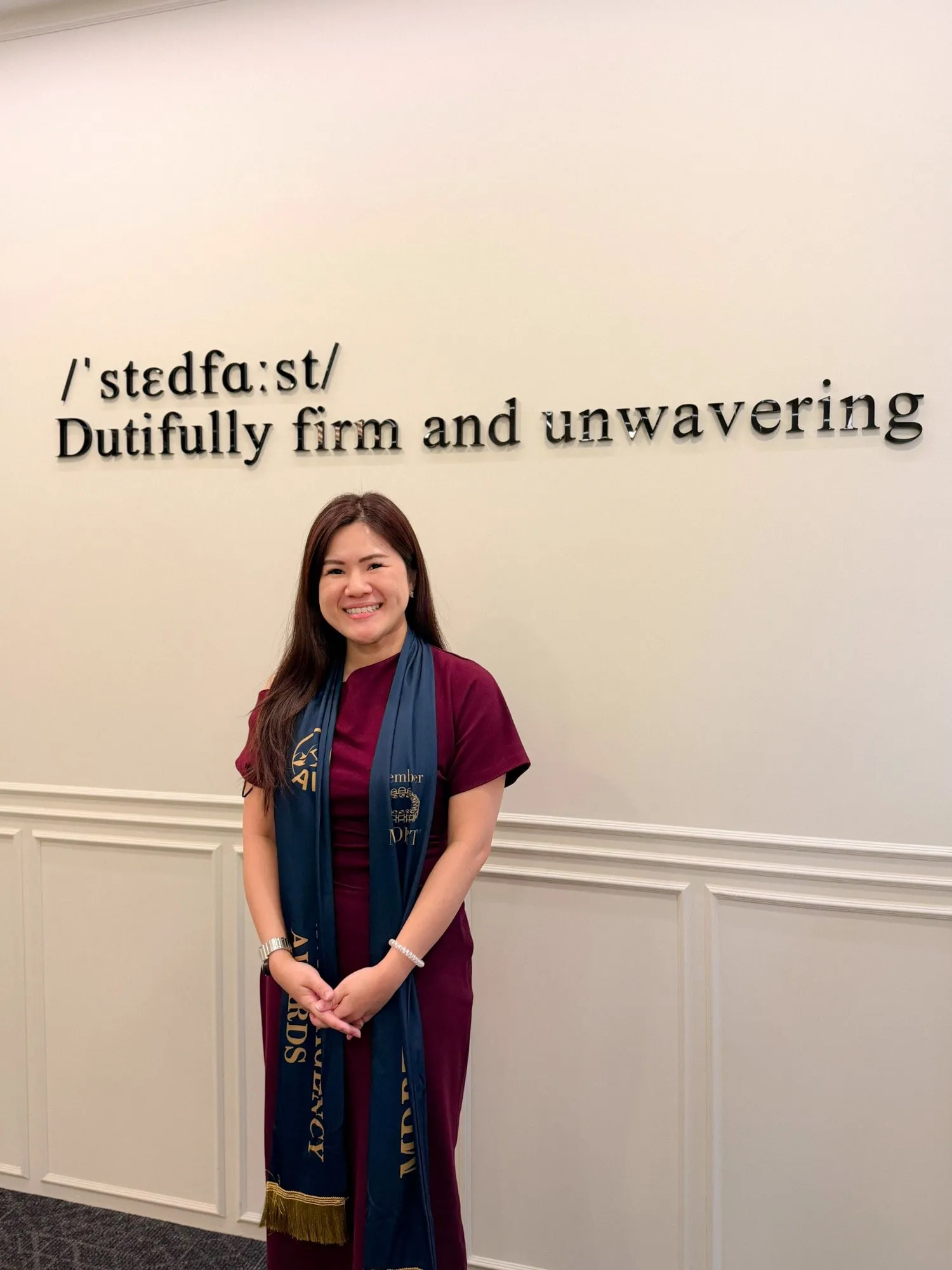 Shermene smiling in a burgundy dress and a navy award sash, standing under a wall sign that reads Dutifully firm and unwavering