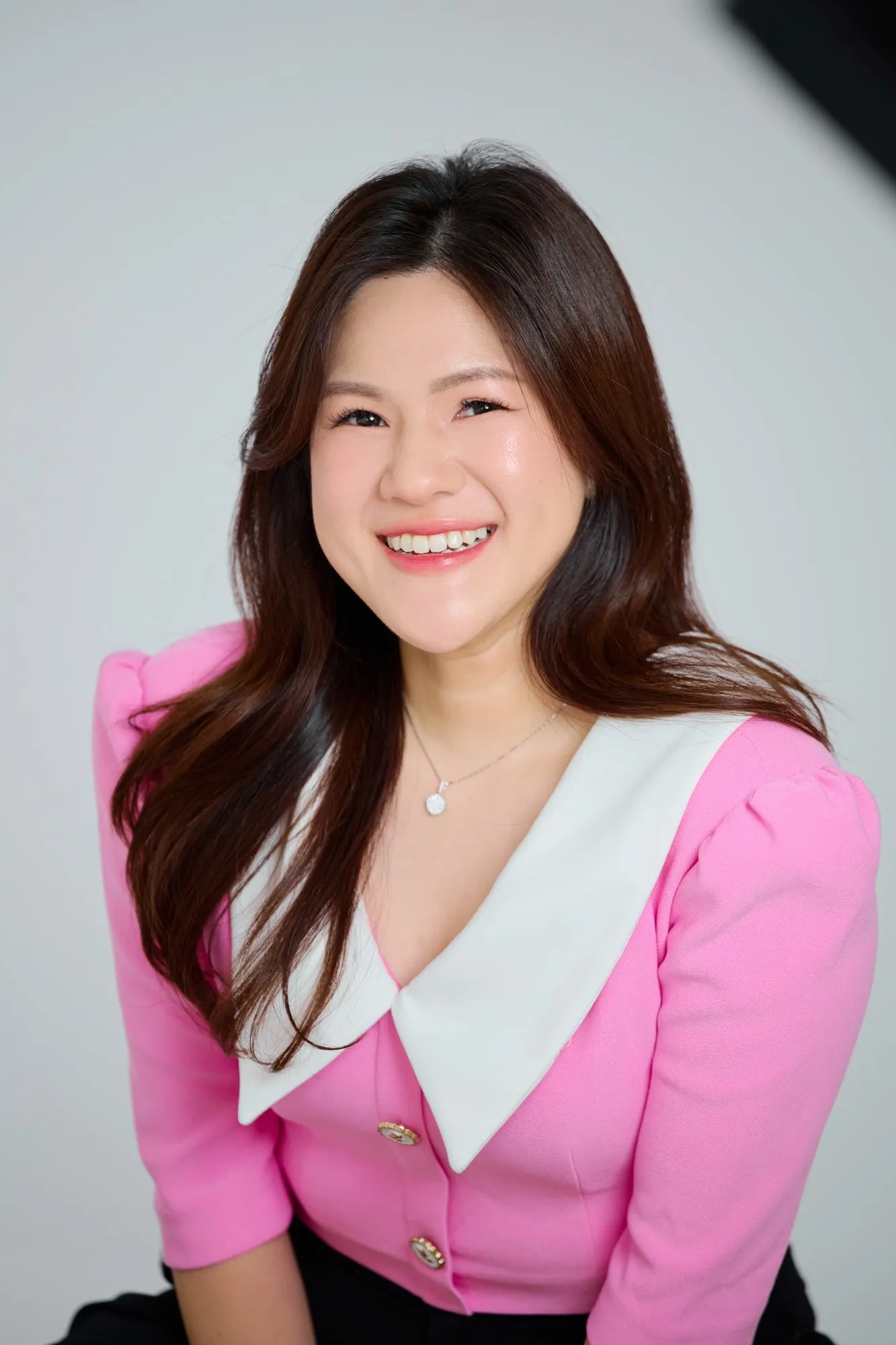 Shermene smiling in a pink jacket with a large white collar, against a soft grey studio background